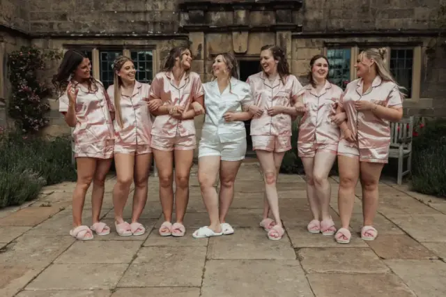 Seven women stand arm-in-arm outdoors at Eyam Hall, smiling and wearing matching satin pajamas and slippers. Six wear pink pajamas, while the bride-to-be in white stands at the center—a joyful moment before weddings begin.