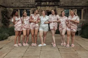 Seven women stand arm-in-arm outdoors at Eyam Hall, smiling and wearing matching satin pajamas and slippers. Six wear pink pajamas, while the bride-to-be in white stands at the center—a joyful moment before weddings begin.
