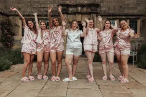 Seven women wearing matching pajamas and slippers stand outdoors at Eyam Hall, smiling and raising their arms in the air. Six wear pink pajamas, while the one in white suggests a fun pre-wedding celebration or bridal party moment together.