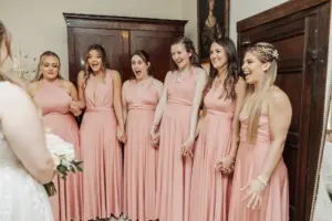 Six bridesmaids in matching pink dresses hold hands and smile with excitement at the bride, who is holding a bouquet and facing them with her back to the camera—capturing pure joy at weddings like those celebrated at Eyam Hall.