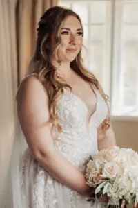 A bride with long wavy hair in a white lace wedding dress holds a bouquet of white and blush roses, standing indoors near a window at Eyam Hall, bathed in soft daylight—perfect for elegant weddings.