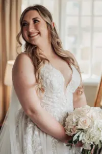 A smiling bride with long, wavy hair holds a bouquet of white flowers. She wears a sleeveless, lace wedding gown and stands indoors at Eyam Hall, bathed in soft natural light—a perfect setting for unforgettable weddings.