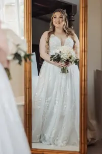 A bride in a white, floral lace wedding dress holds a bouquet and smiles while looking at her reflection in a large, gold-framed mirror at historic Eyam Hall—capturing the timeless charm of weddings.