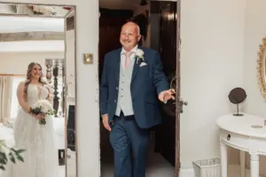 A smiling man in a blue suit enters a room at Eyam Hall, reacting with joy as he sees a bride in a white dress holding a bouquet, standing near a mirror and white vanity—capturing the magic of weddings.