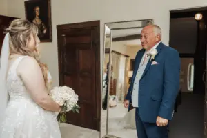 A bride in a white dress holds a bouquet and faces an emotional, teary-eyed older man in a blue suit. They are indoors at Eyam Hall, near a large mirror and dark wooden doors, capturing the heartfelt moments weddings bring.