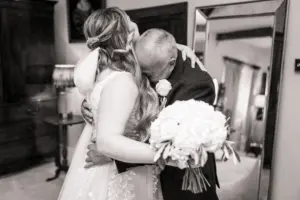 A bride in a wedding dress hugs an older man tightly, both emotional. The bride holds a bouquet of white flowers. They stand in a warmly lit room at Eyam Hall, with a large mirror and wooden furniture—capturing the warmth of weddings.