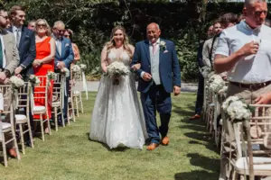 A smiling bride in a white gown walks down an outdoor aisle at Eyam Hall with an older man in a suit, surrounded by standing wedding guests on a sunny day.