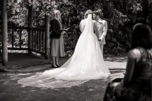 A bride and groom stand facing each other during an outdoor Eyam Hall wedding ceremony, with the bride in a flowing gown and veil. An officiant stands nearby, and a guest is seated in the foreground amid lush, sunny surroundings.