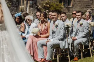 Wedding guests sit outdoors at Eyam Hall, dressed in formal attire. A woman in a pink dress holds flowers, while men in light gray suits sit beside her. The sunny setting features weddings charm with a stone building and greenery in the background.