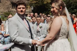 Guests seated outdoors at Eyam Hall watch a beautiful wedding ceremony, the focus on their faces as the bride and groom stand holding hands, slightly out of focus in the foreground.