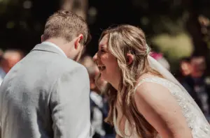 A bride in a white dress and veil laughs joyfully next to a groom in a light grey suit during an outdoor wedding ceremony at Eyam Hall. The background is softly blurred with guests and greenery visible, capturing the magic of weddings.