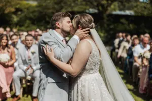 A bride and groom share a kiss during their outdoor Eyam Hall wedding ceremony, surrounded by seated guests on a sunny day. The bride wears a white lacy gown and veil; the groom is in a light gray suit.