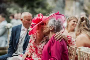 Two older women in bright pink outfits and hats kiss affectionately at an outdoor wedding at Eyam Hall, surrounded by smiling guests sitting on chairs. The mood is joyful and celebratory—capturing love and happiness often found at weddings.