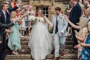 A bride and groom smile as they walk down the outdoor steps of Eyam Hall, holding hands, while guests cheer and toss flower petals around them at their weddings celebration.