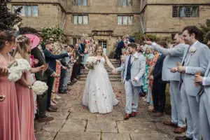 A bride and groom walk hand in hand down a stone path at Eyam Hall, smiling as wedding guests in colorful attire shower them with rose petals outside the historic stone building.