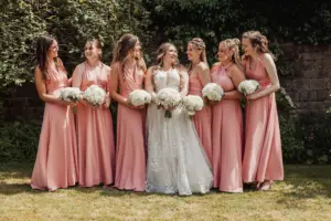 A bride in a white dress stands outside Eyam Hall with six bridesmaids in matching pink dresses, all holding white bouquets and smiling at each other. They are on grass with a stone wall and green foliage, ready to celebrate weddings together.