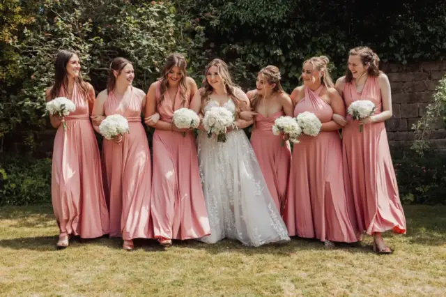A bride in a white gown stands in the center of six bridesmaids wearing matching blush pink dresses at Eyam Hall, all smiling and holding white bouquets outdoors on a sunny weddings day.