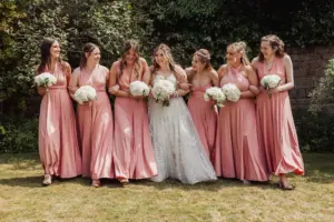 A bride in a white gown stands in the center of six bridesmaids wearing matching blush pink dresses at Eyam Hall, all smiling and holding white bouquets outdoors on a sunny weddings day.