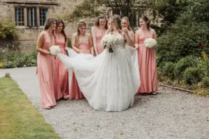 A bride in a white gown is surrounded by six bridesmaids in matching pink dresses, holding white bouquets and helping arrange her dress as they walk outside Eyam Hall on a gravel path near stone buildings and lush greenery—perfect for weddings.