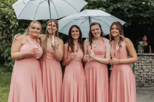 Five women in matching blush pink dresses stand outdoors at Eyam Hall weddings, smiling and holding glasses of champagne. Two hold white umbrellas, with green foliage and a stone wall in the background.