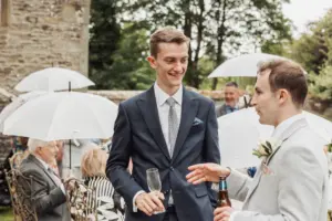 Two men in suits talk and smile at an outdoor Eyam Hall wedding, holding drinks. People sit behind them under white umbrellas, with greenery and a stone building in the background.