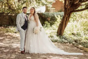 A bride in a white gown and veil stands smiling beside a groom in a light gray suit and tie, holding a bouquet of flowers at their Eyam Hall wedding, surrounded by trees and greenery in a sunlit garden.