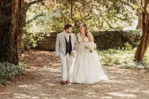 A bride and groom walk hand-in-hand outdoors at Eyam Hall, smiling at each other. The bride wears a white gown and holds a bouquet, while the groom wears a light gray suit. Sunlight filters through the surrounding trees, capturing the magic of weddings.