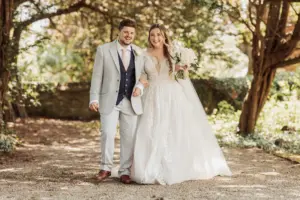 A smiling bride in a white gown holding a bouquet stands next to a groom in a light gray suit and navy vest. They hold hands and walk outdoors under sunlit trees at Eyam Hall, surrounded by greenery—a perfect moment for weddings.