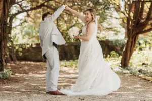 A bride in a white dress holding a bouquet smiles as her partner in a light suit twirls her under sunlit trees, capturing the joy of weddings at Eyam Hall.