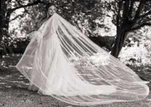 A bride in a flowing gown stands outdoors at Eyam Hall, smiling and holding her bouquet, as her long, sheer veil billows gracefully to the side. The scene is shaded by trees and captured in black and white, evoking timeless weddings elegance.