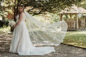 A bride in a white wedding dress stands outdoors at Eyam Hall, holding a bouquet of flowers. Her long veil flows behind her, and she smiles under the shade of a tree with a wooden gazebo in the background, capturing the magic of weddings.
