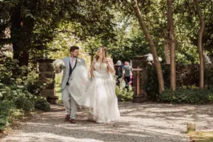 A bride and groom walk together outdoors on a sunny day at Eyam Hall, smiling and holding hands as the groom carries the bride’s bouquet. They are surrounded by greenery, trees, and wedding guests visible in the background.