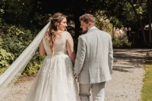 A bride and groom walk hand-in-hand along a sunlit garden path at Eyam Hall, looking at each other and smiling. Surrounded by greenery, their wedding day is picture-perfect—the bride in a white dress with a long veil, the groom in a light gray suit.
