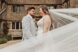 A bride and groom stand facing each other outdoors at Eyam Hall, holding hands and smiling. The brides long veil flows in the breeze, creating a magical moment in front of the historic stone building’s large windows—a perfect setting for weddings.