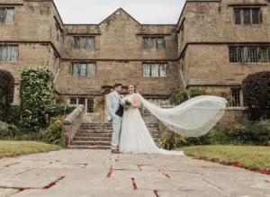 A bride and groom stand together in front of historic Eyam Hall, her long veil flowing in the wind. Dressed in wedding attire, they are surrounded by lush greenery and beautiful gardens, capturing the essence of unforgettable weddings.