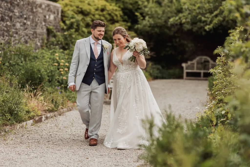 A bride and groom walk together along a garden path at Eyam Hall, smiling. The bride wears a white lace gown and holds a bouquet, while the groom wears a light grey suit with a navy vest. Greenery and stone walls surround them—pure weddings bliss.