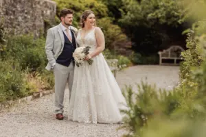 A bride in a white lace gown holding a bouquet stands beside a groom in a light grey suit and navy vest on a gravel path in the gardens of Eyam Hall, both smiling and looking to their left, capturing the joy of weddings.