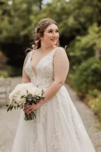 A smiling bride in a sleeveless, lace wedding gown holds a bouquet of white roses and greenery while standing outdoors on a garden path at Eyam Hall, the perfect setting for unforgettable weddings.