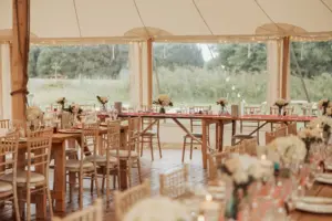 A rustic weddings reception setup in a bright tent at Eyam Hall features wooden chairs and tables arranged in rows, adorned with white flowers, candles, and tableware. Large windows reveal beautiful greenery outside.