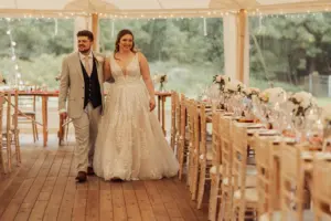 A smiling bride and groom walk hand in hand inside a decorated tent at Eyam Hall weddings, with wooden floors, floral centerpieces, set tables, and string lights twinkling amid lush greenery outside.