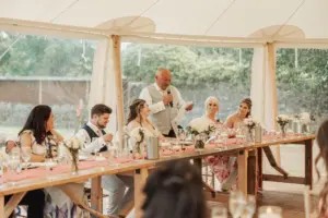 A man stands and speaks into a microphone at a long, decorated table during an Eyam Hall wedding reception, surrounded by guests who are seated and listening attentively under a bright tent.