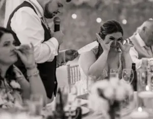 A bride wipes away tears while seated at a table during a heartfelt speech at Eyam Hall weddings, as a man stands beside her speaking into a microphone. Other guests also appear emotional during the touching moment.