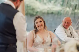 A bride in a white dress smiles warmly at a man giving a speech, while an older man in a vest and tie sits beside her at a decorated table during weddings at Eyam Hall.