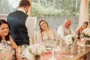A bride smiles lovingly at the groom giving a speech during a joyful wedding reception at Eyam Hall. Guests, including an older man and two women, sit at a decorated table adorned with flowers and drinks, celebrating weddings in style.