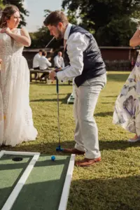A man in formal attire plays mini golf outdoors beside two women in dresses at Eyam Hall. The sunny scene suggests a festive atmosphere, perfect for weddings, with guests seated at a picnic table in the background.