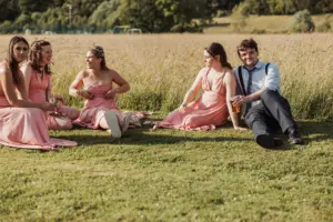 Five people sit on grass by a field at Eyam Hall, four women in pink dresses and one man in a white shirt with suspenders. They appear relaxed, chatting and smiling with drinks in hand—capturing the joy of weddings on a sunny day.
