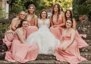 A bride in a white gown sits on stone steps at Eyam Hall, surrounded by six bridesmaids in matching pink dresses, all smiling and posing outdoors with lush greenery—capturing the joy of weddings in a charming setting.