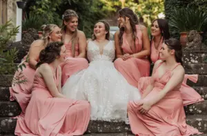 A bride in a white gown sits on outdoor steps at Eyam Hall, surrounded by six bridesmaids in matching pink dresses, all smiling and laughing together amidst the beautiful garden—capturing the joyful spirit of weddings.