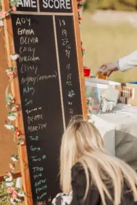 At Eyam Hall, a woman with blonde hair writes on a large chalkboard scoreboard decorated with artificial flowers—perfect for weddings. The board lists names and scores, while a picnic table with snacks and drinks is set up beside her outdoors.