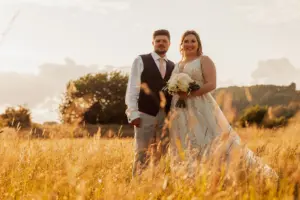 A bride and groom stand together in a sunlit field near Eyam Hall, surrounded by tall grass and trees, with the bride holding a bouquet of flowers—both smiling joyfully at the camera, capturing the magic of weddings.